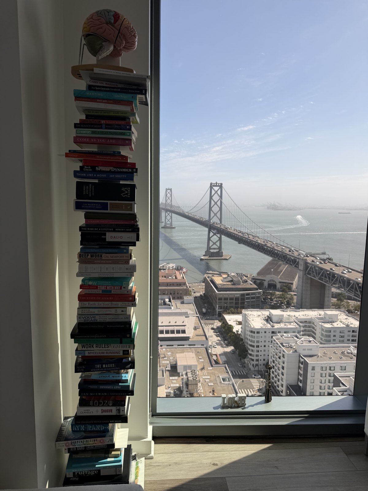 A floor-to-ceiling tower of stacked books beside a window overlooking the Bay Bridge in San Francisco
